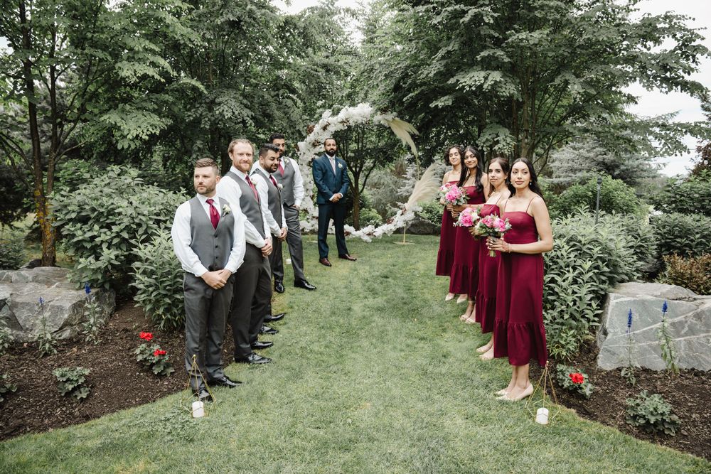 Wedding party lined up outdoors on garden path with bridesmaids in burgundy dresses and groomsmen in dark suits.