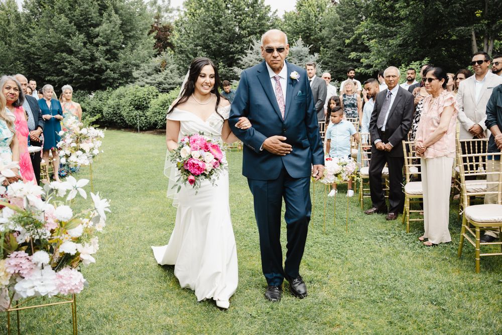 Wedding processional on manicured lawn with pink floral arrangements and gold chairs.