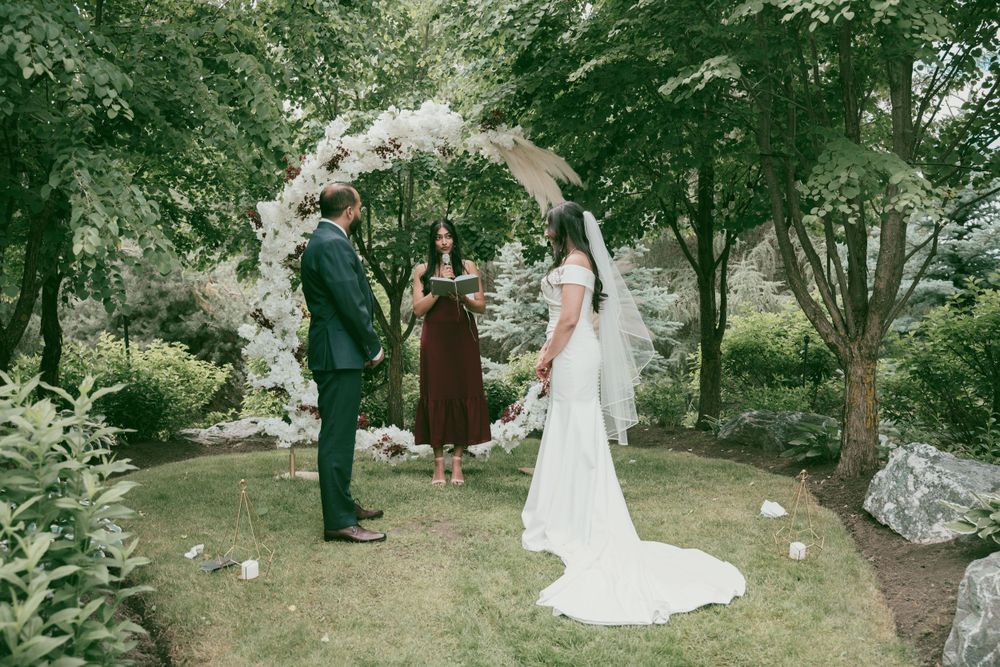 Outdoor wedding ceremony under floral arch with couple and officiant surrounded by greenery.