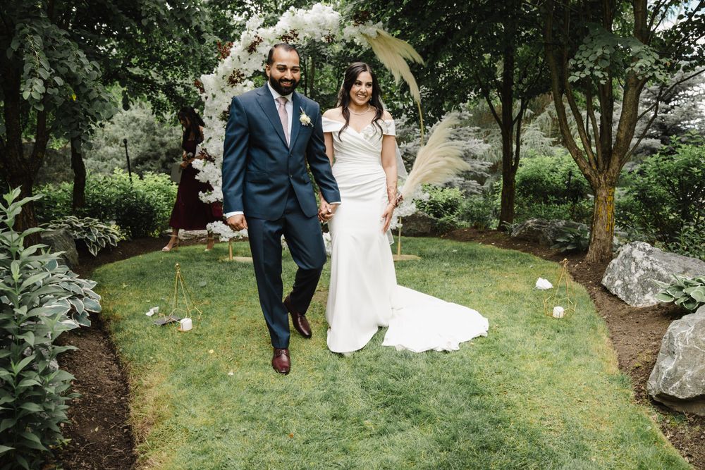 Newlyweds pose together under white floral arch in garden setting.