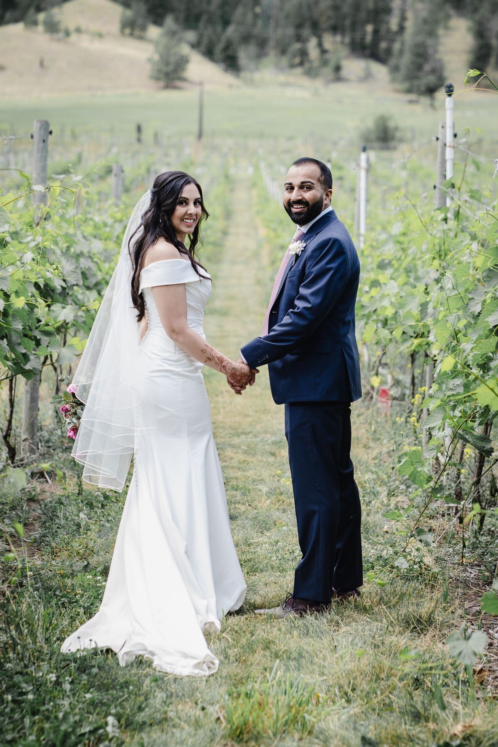 Couple poses in vineyard wearing formal wedding attire with mountain backdrop.