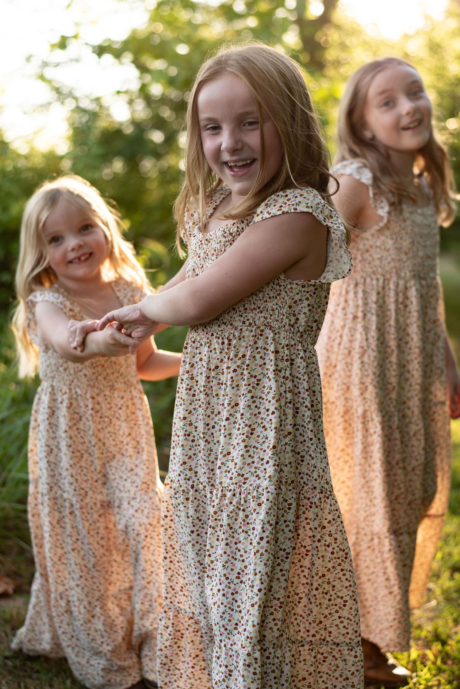 Three young girls in matching floral dresses dancing in golden sunlight.