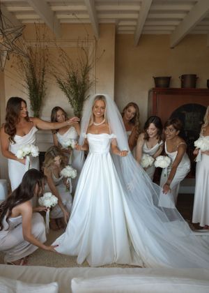 Wedding party scene with bridesmaids in white dresses surrounding bride in elegant ballgown.