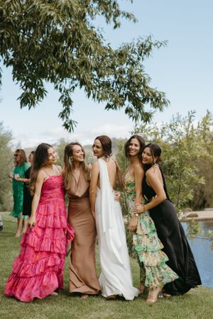 Bridesmaids in colorful dresses posing together by a lake on a sunny day.