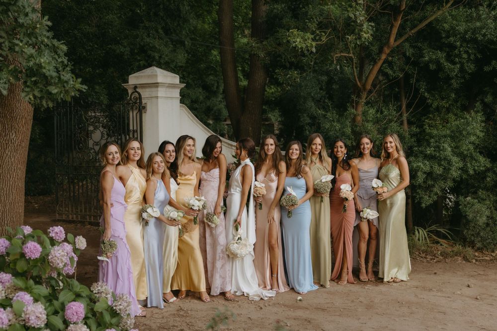 Wedding party members in pastel and neutral dresses stand in a row against a wooded backdrop.