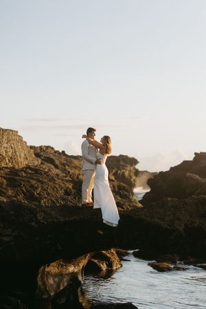 A couple in wedding attire embraces on rocky coastal cliffs at sunset near crashing waves.