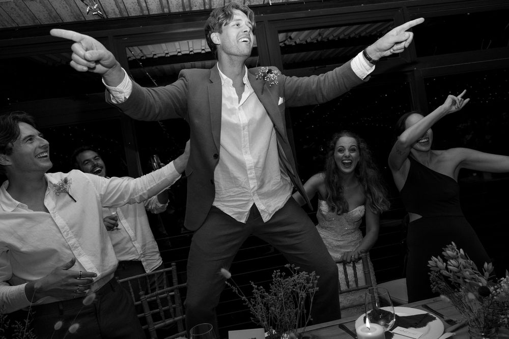 Wedding reception guests dancing and celebrating with arms raised in a dark indoor venue.