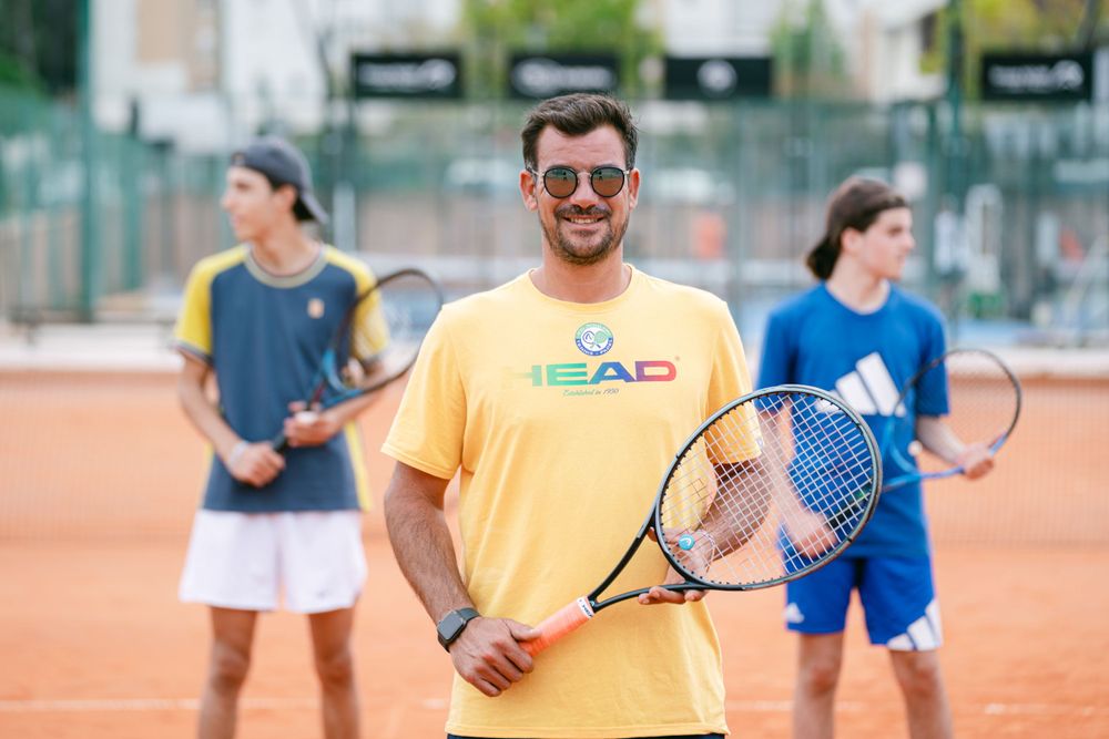 Tennis coach in bright yellow shirt smiling on clay court during outdoor lesson.