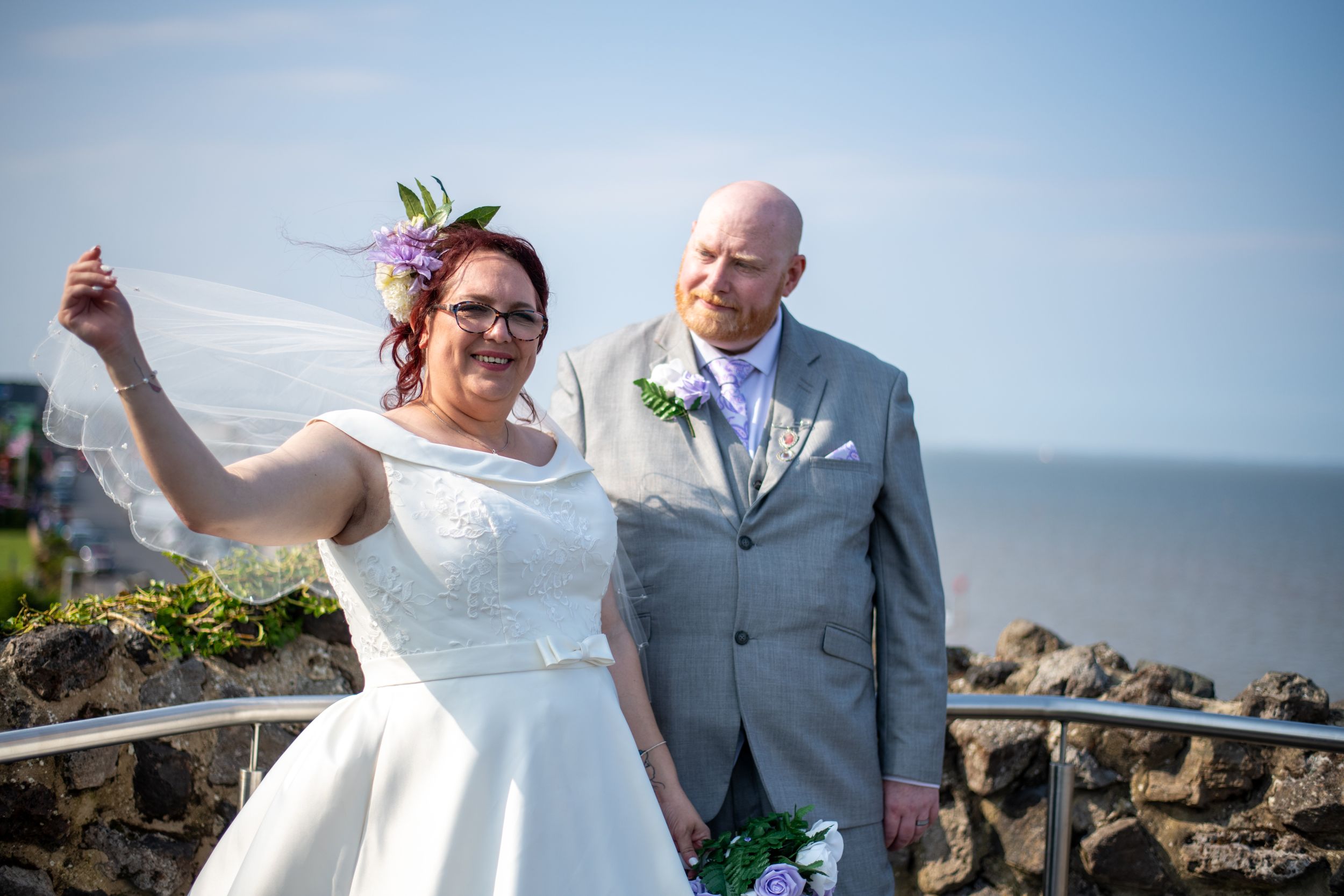Joyful wedding couple posing by seaside stone wall under blue skies.