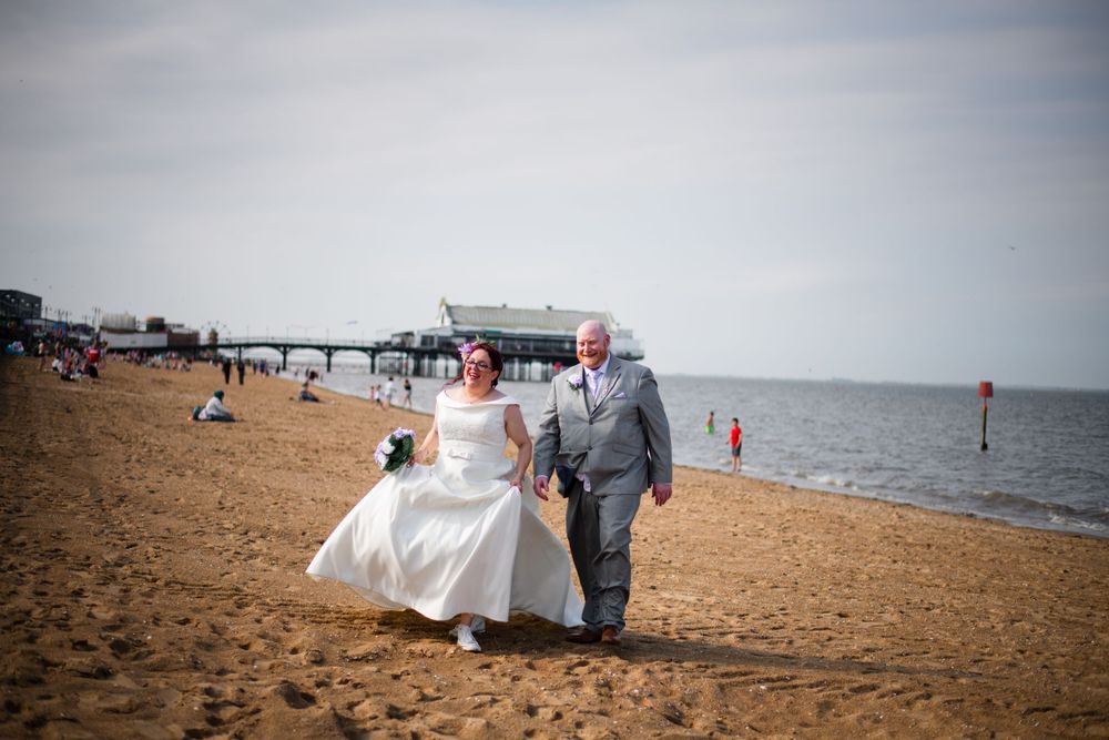 A couple walks together on a sandy beach with a white wedding dress flowing in the breeze.