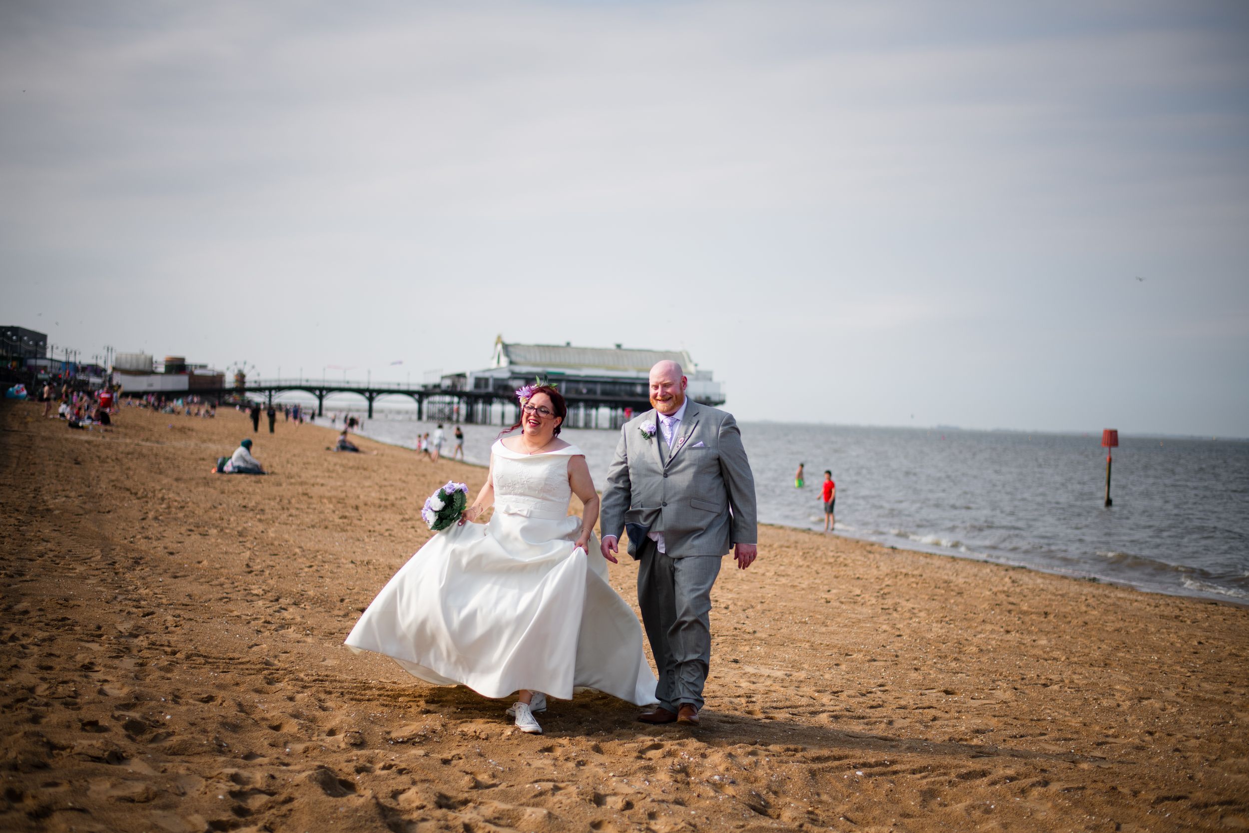 A couple walks together on a sandy beach with a white wedding dress flowing in the breeze.