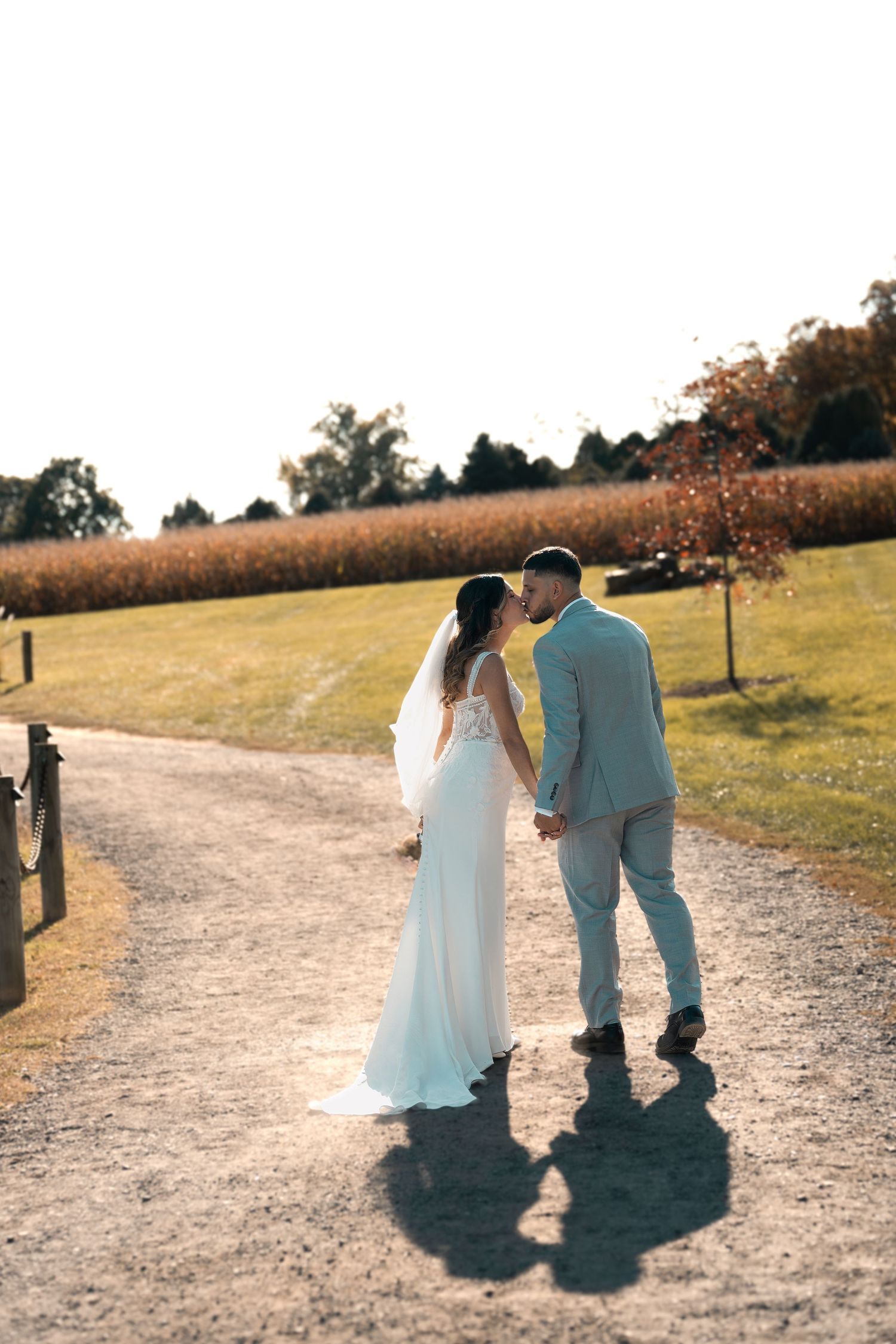 Newlywed couple walking down a sunlit path with cornfields and autumn trees in the background on their wedding day.