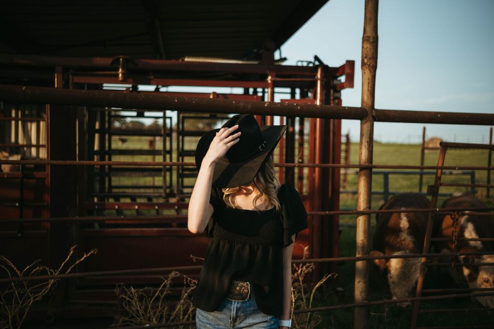Person in black adjusts hat while standing near metal livestock gates at sunset.
