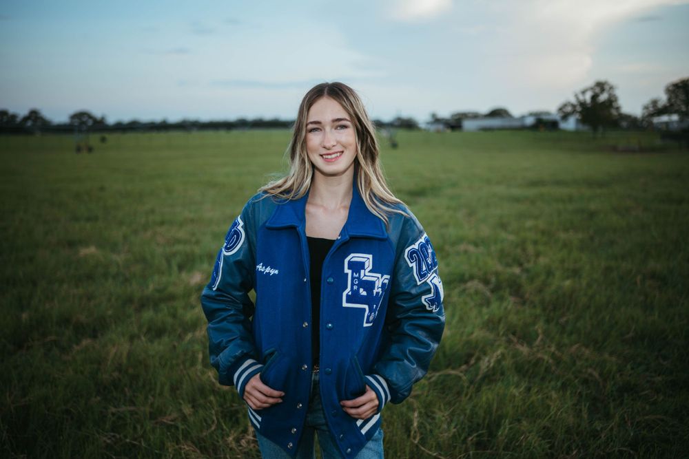 Blue varsity jacket with letter patches worn in grassy field.