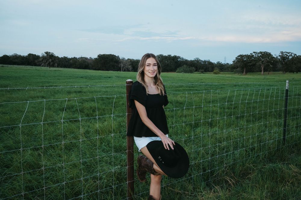 A person in casual attire poses by a wire fence in a grassy field with a black cowboy hat.