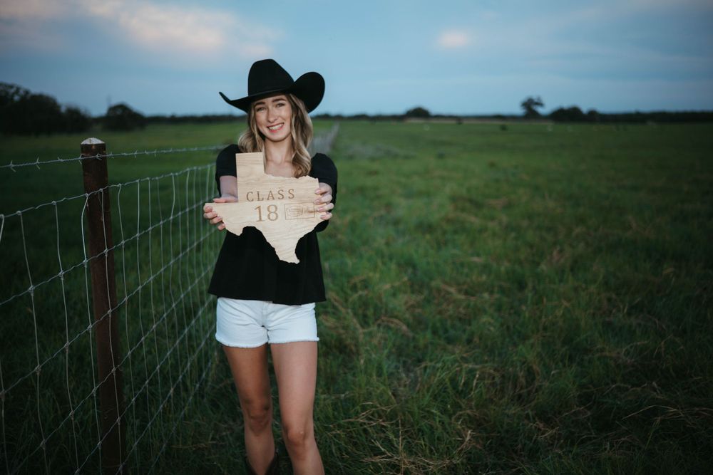 A figure in a black cowboy hat and shorts stands by a wire fence in a rural field holding a sign.