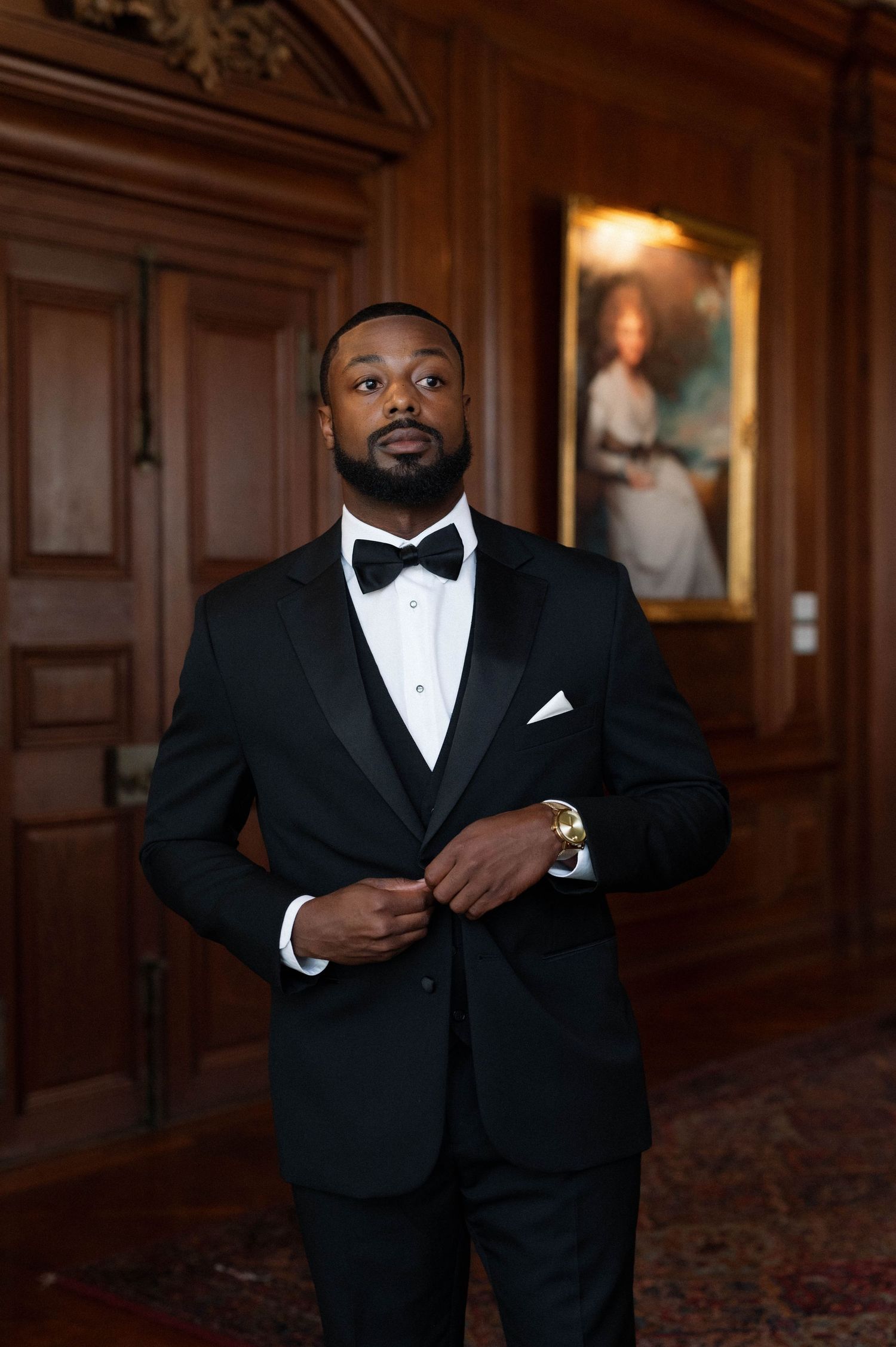 Groom in black bow tie and tuxedo standing in elegant wood-paneled hallway.