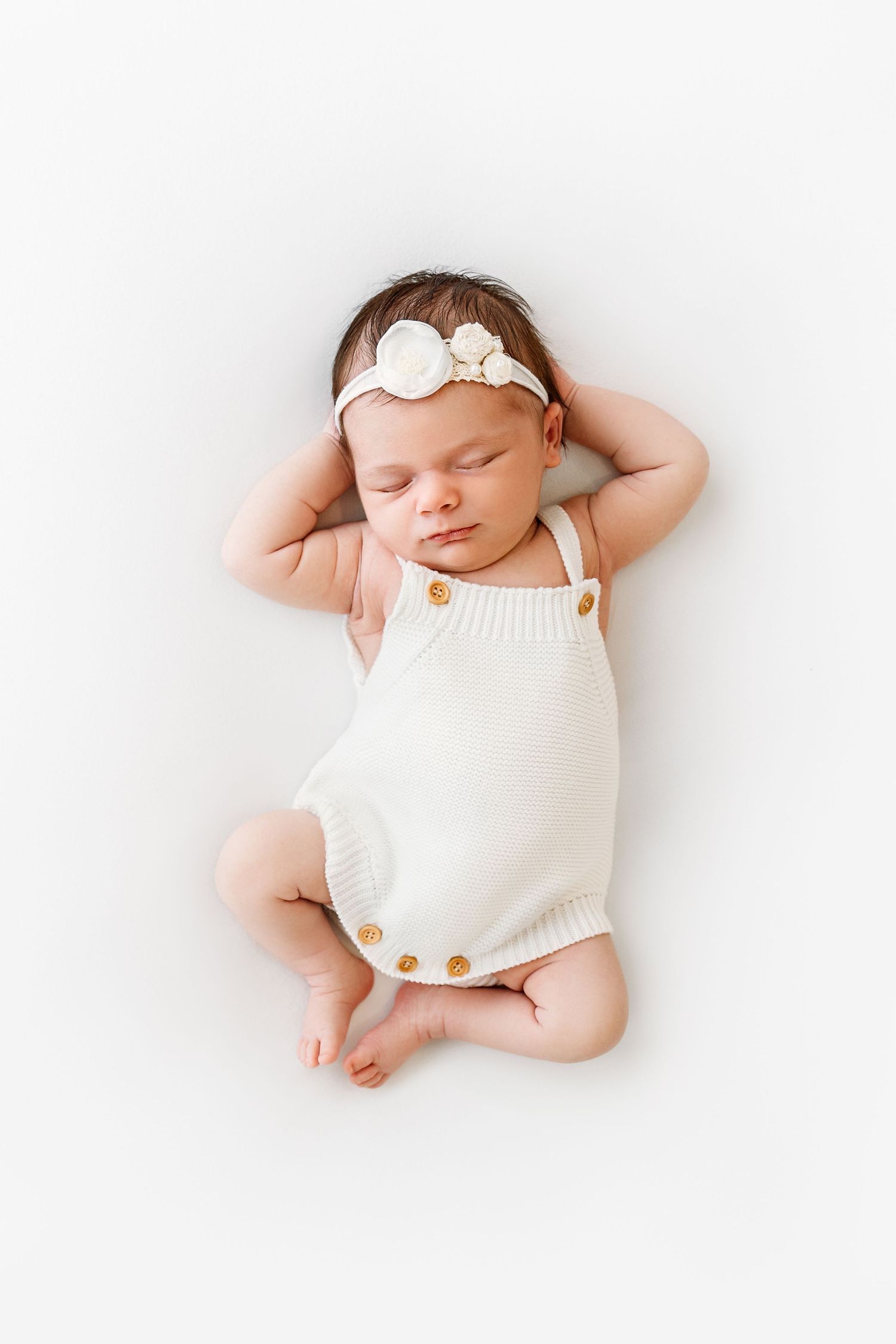 Newborn infant girl in knit white onesie, looking relaxed with hands behind head and legs looking similar