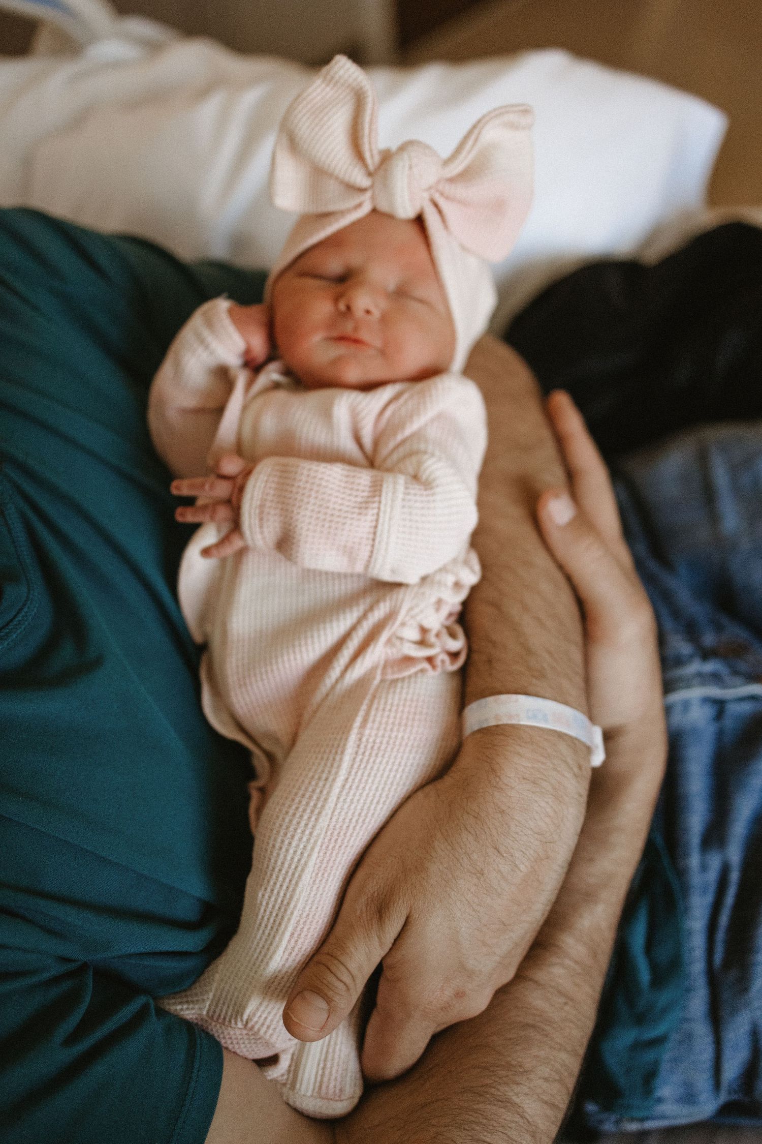Sleeping baby wearing soft pink bodysuit with matching bow headband rests peacefully on adult's lap with teal blanket.