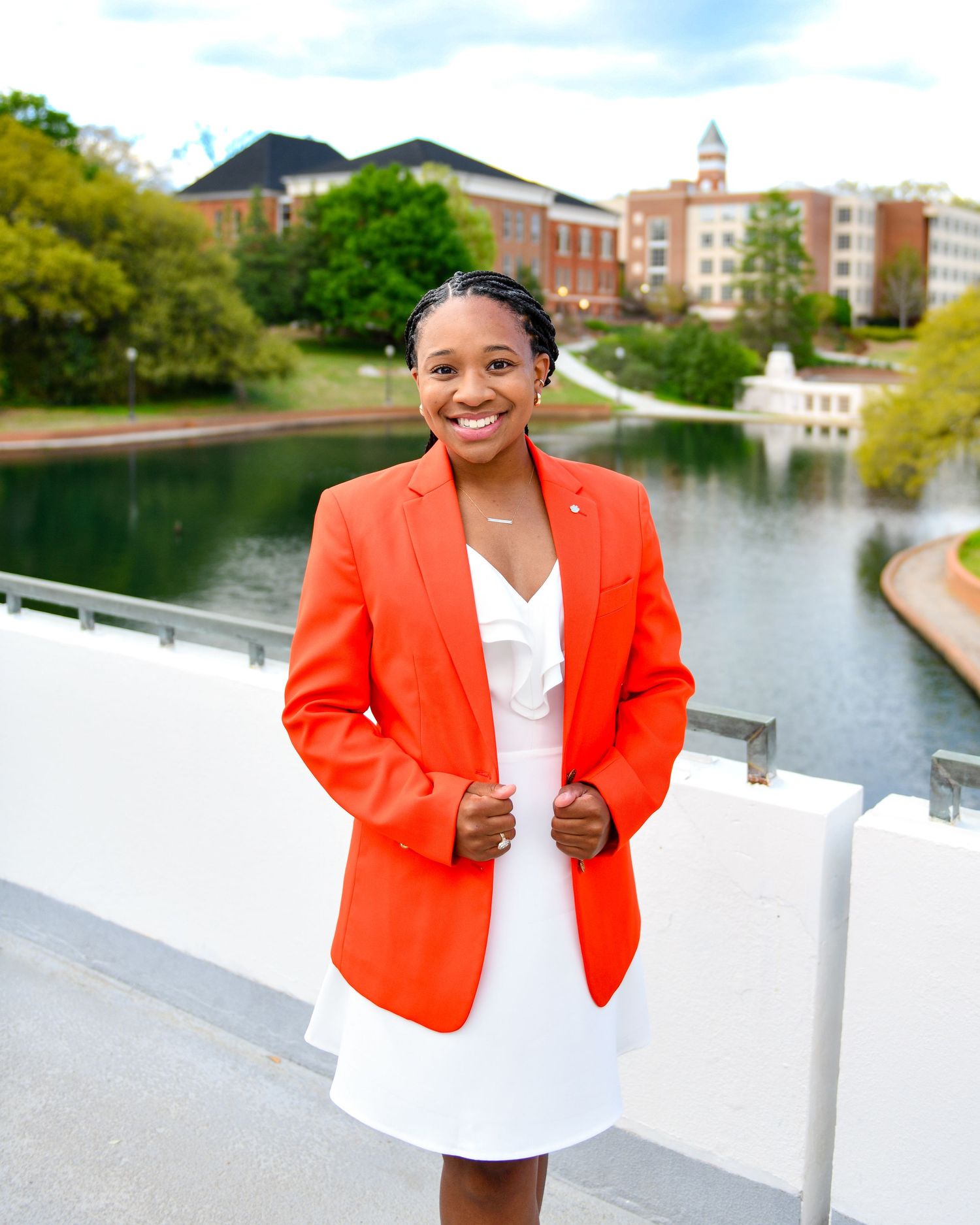 Professional in bright orange blazer and white dress poses by scenic campus lake.