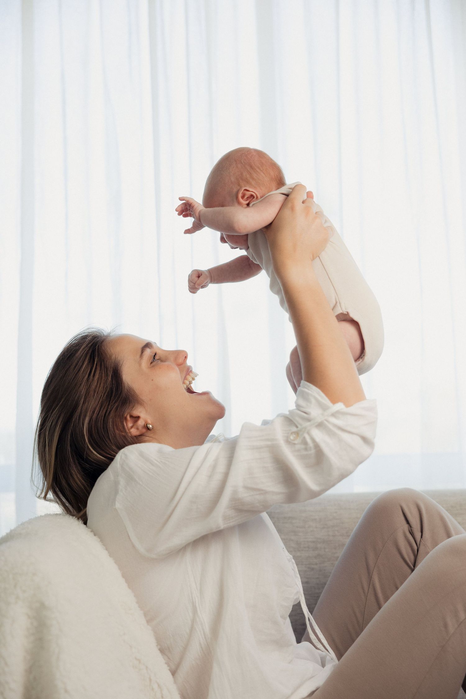 A joyful mother lifts her newborn baby above her head while sitting on a couch near bright white curtains.