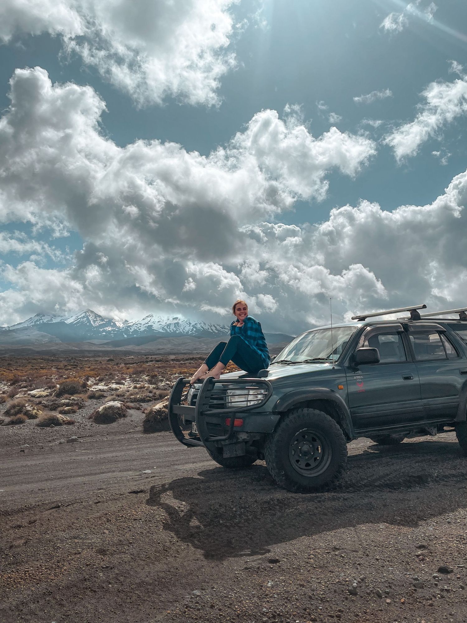 Person sitting on off-road vehicle in desert landscape with snow-capped mountains and dramatic clouds.