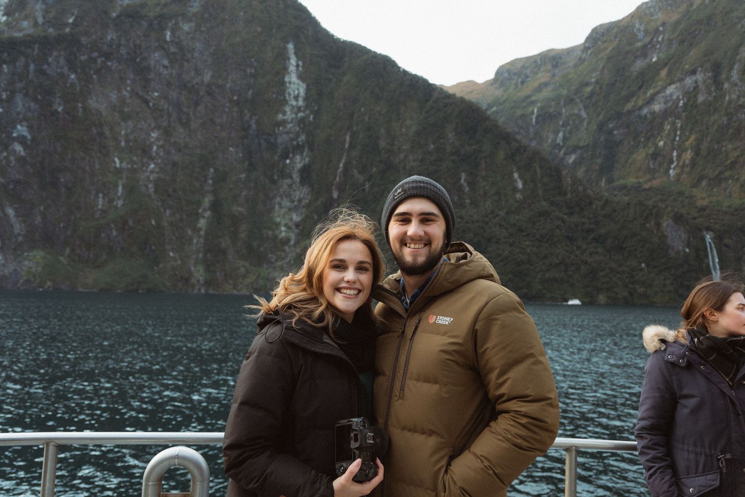 Couple smiling on a boat tour at Milford Sound, New Zealand, with dramatic fjord cliffs and waterfalls behind them.