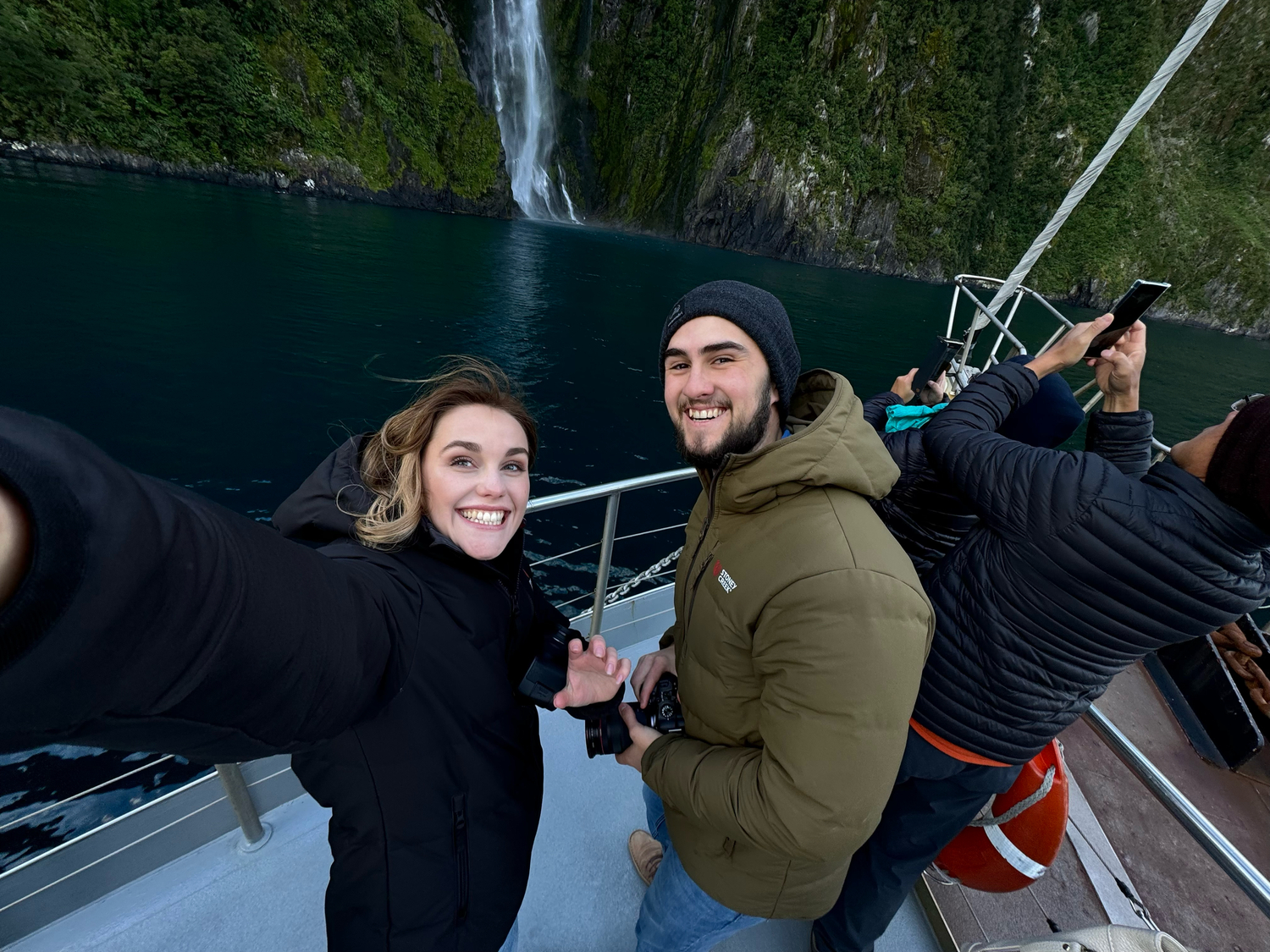 Couple taking selfie on boat with dramatic waterfall backdrop.