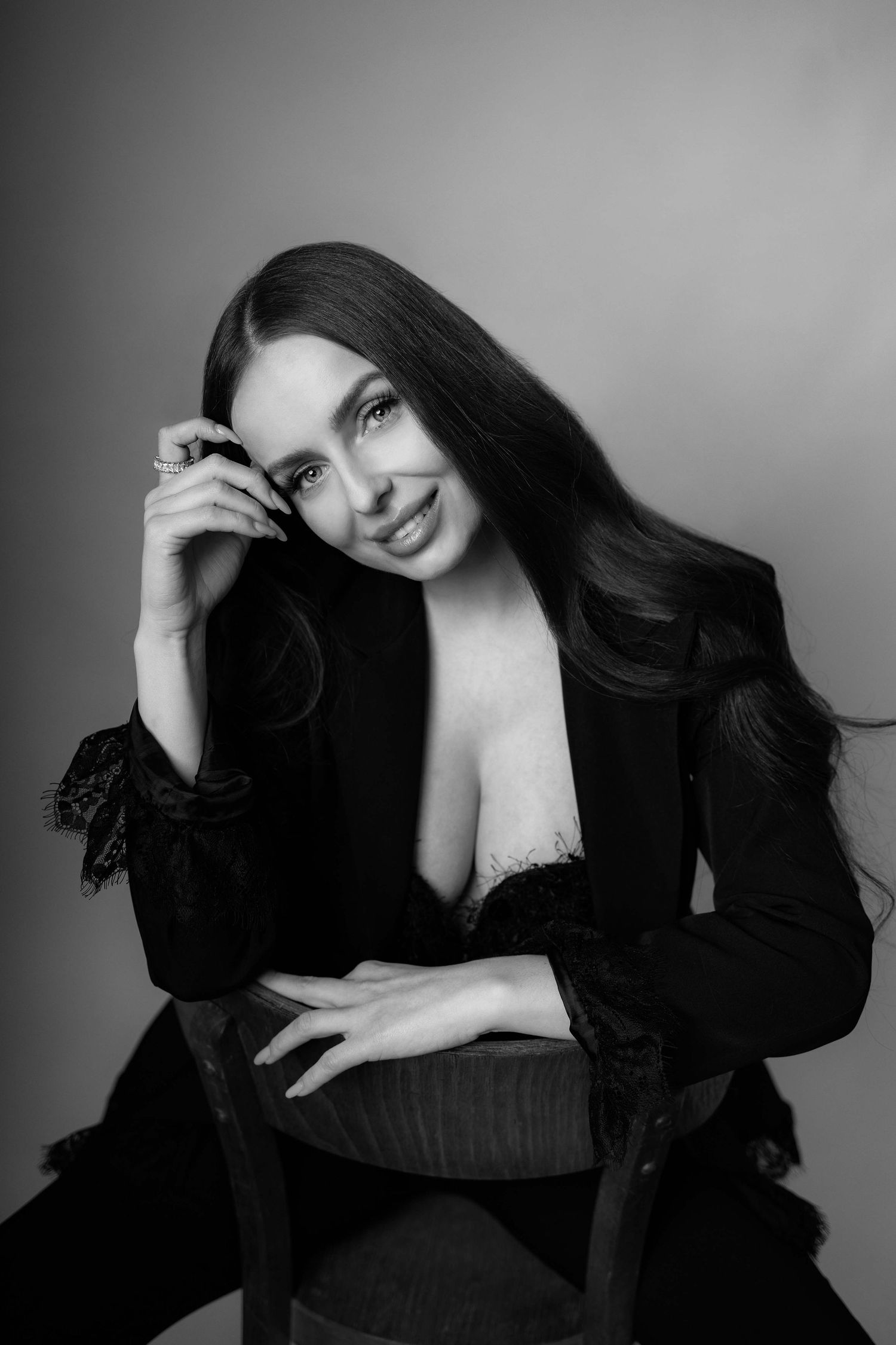Čiernobiely štúdiový portrét usmiatej brunetky v čiernom čipkovanom saku sediacej na drevenej stoličke. Svadobný fotograf, Rajecká dolina.