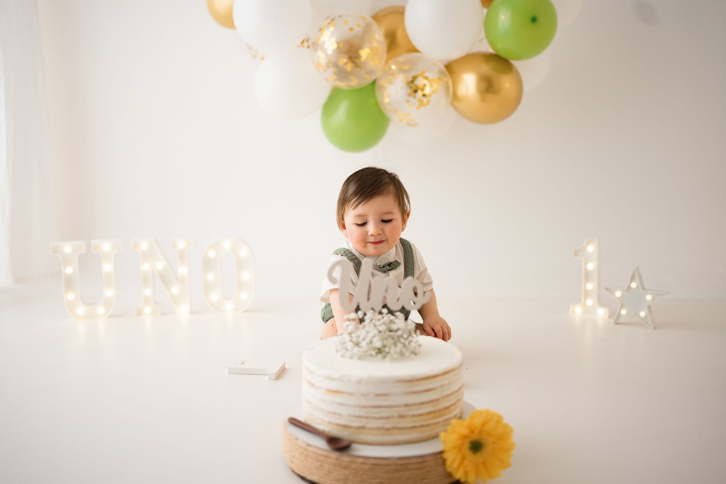 Baby boy in green overalls sits before a white layered cake at a first birthday smash cake photo session.