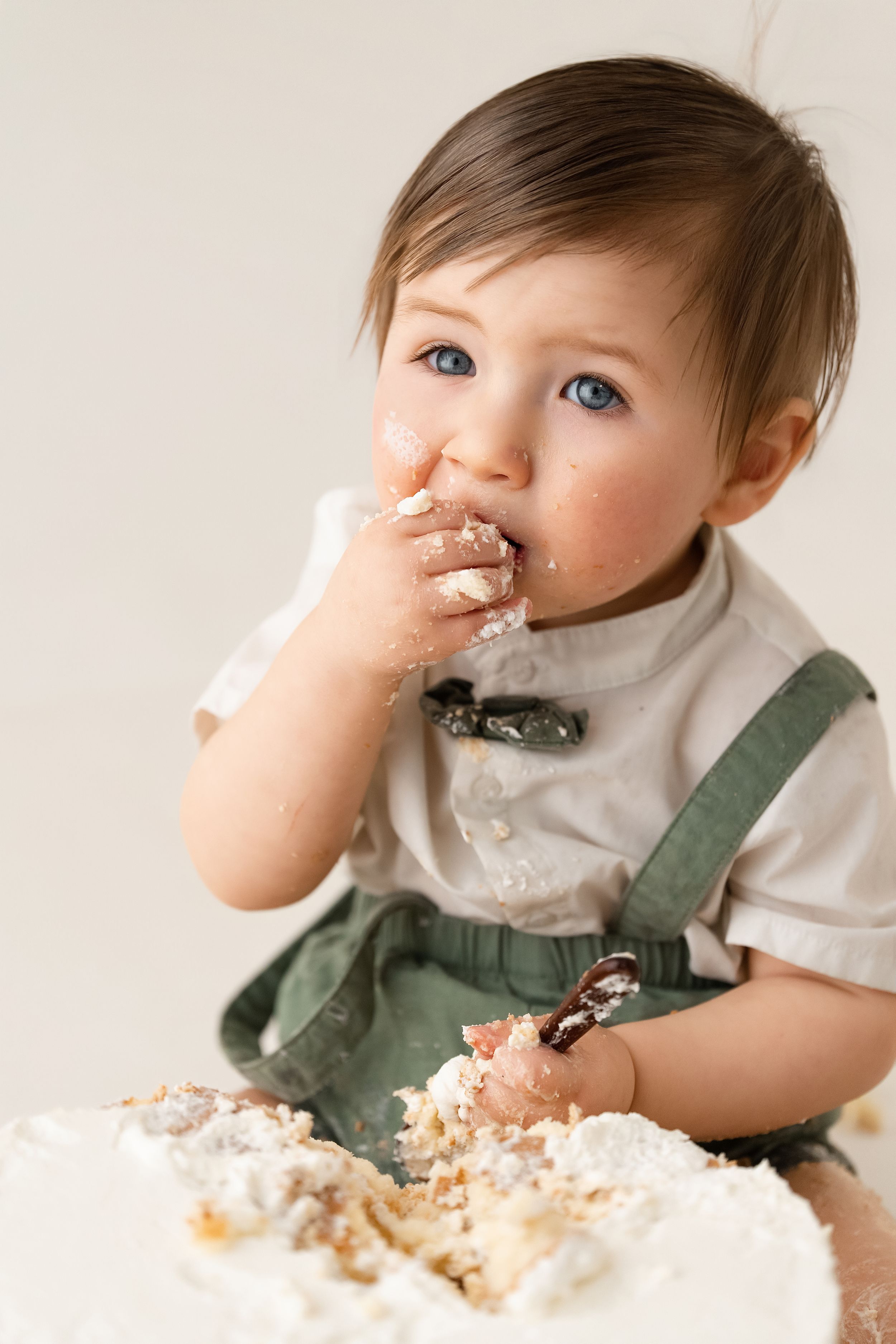 Baby boy in green overalls eagerly eats birthday cake with both hands during a cake smash session.