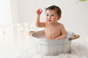 Happy baby boy splashes in a galvanized tub bath after a first birthday cake smash photo session.