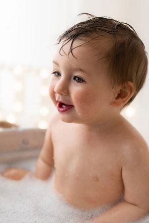 Smiling baby boy with wet hair enjoys a bubbly bath after a first birthday cake smash celebration.
