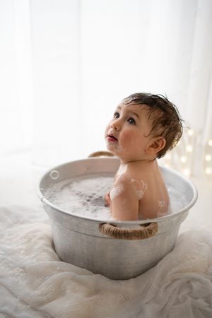 A wet toddler sits in a vintage metal wash tub filled with soapy bubbles during a bath photo session.