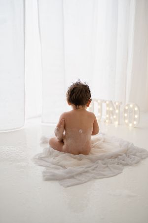 Rear view of a wet naked toddler sitting on white fabric after a bath in a bright white studio.
