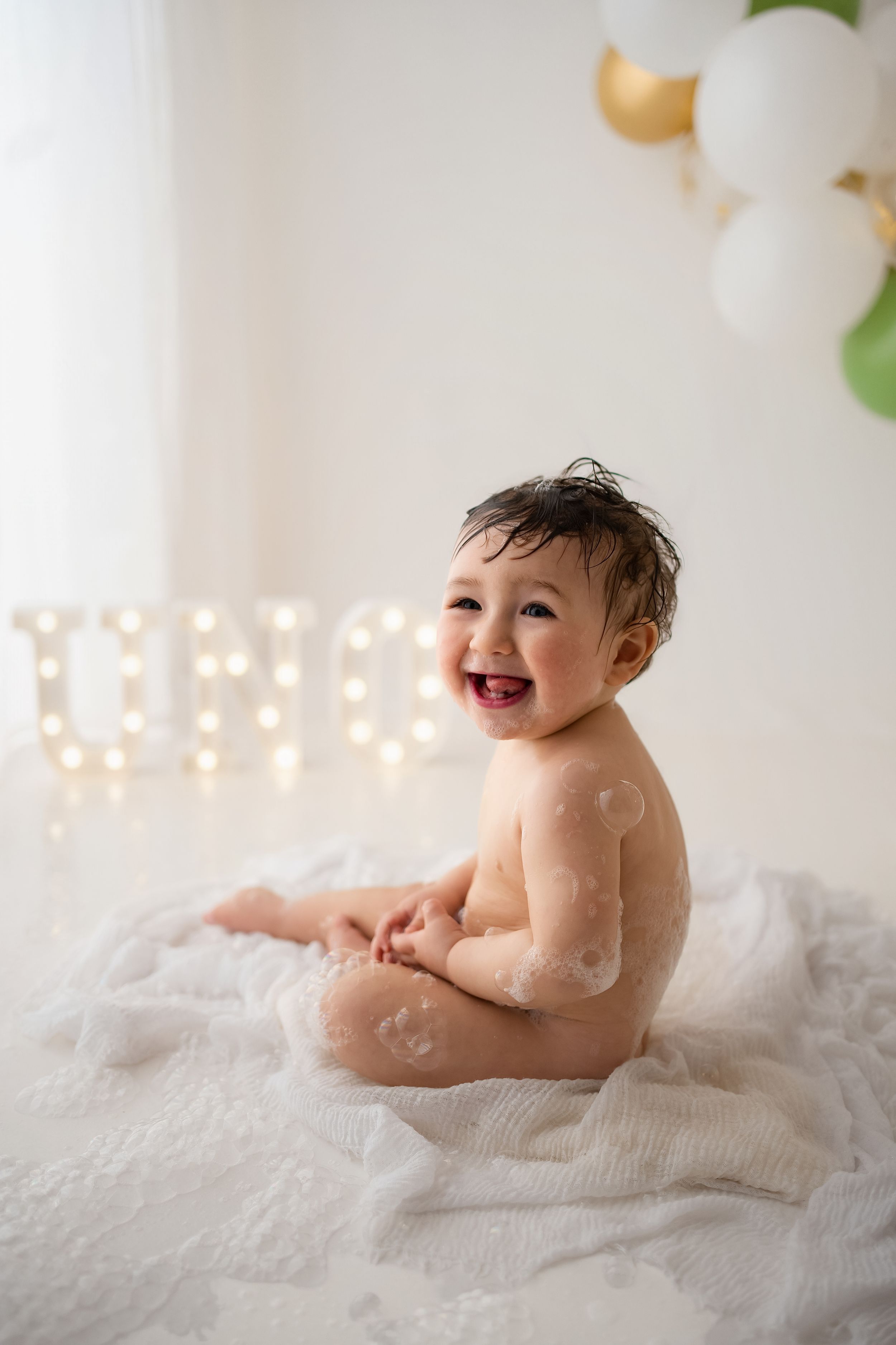 Happy laughing baby covered in soap suds sits on white blanket near illuminated ONE sign with balloons.