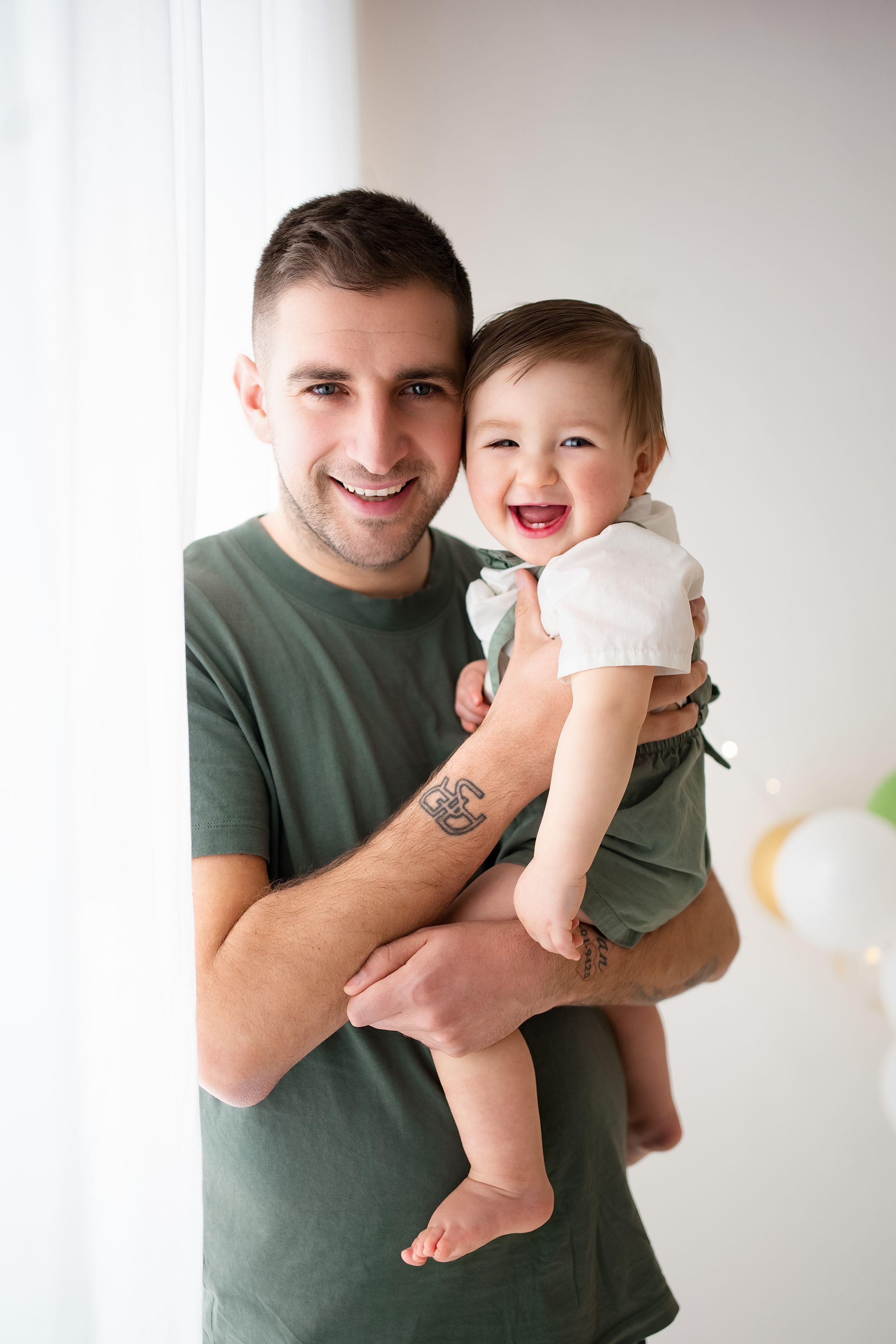 A smiling father in a green t-shirt holds his laughing baby girl near a bright window.