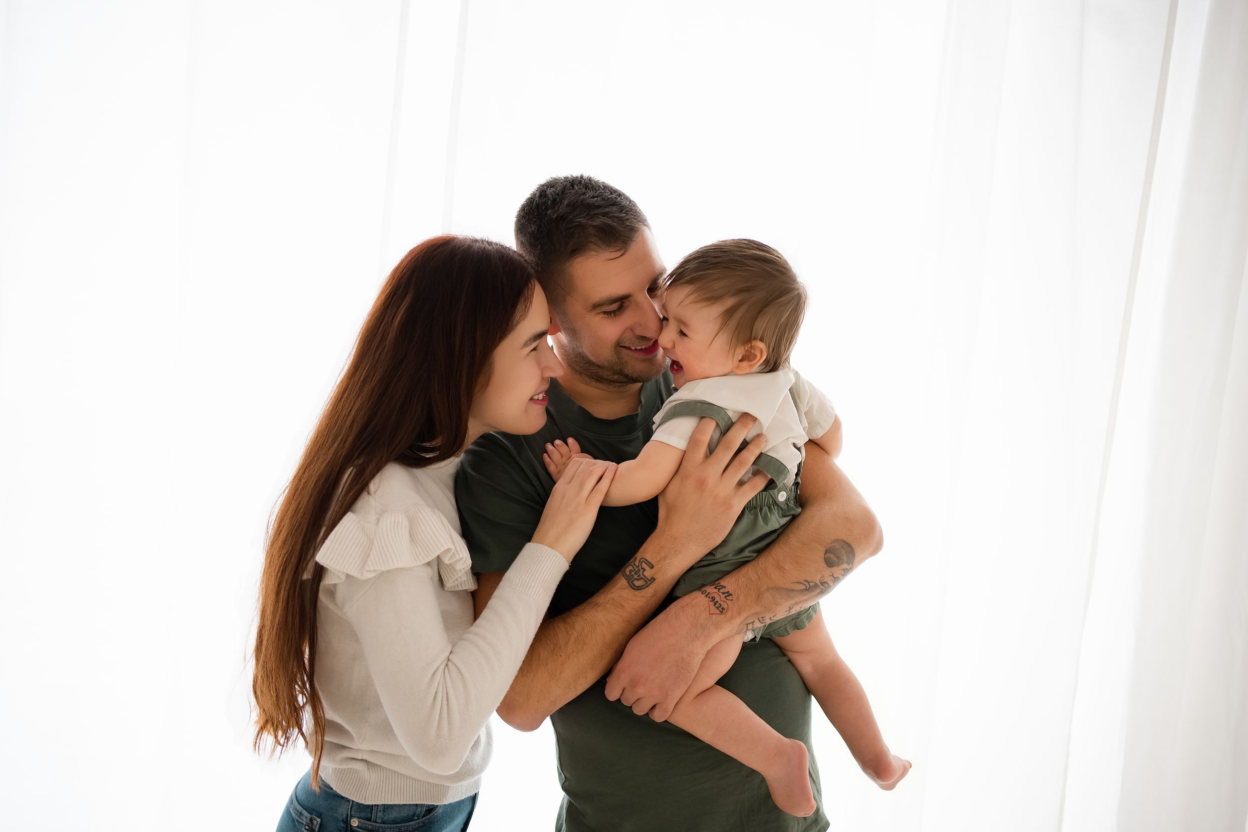 A loving couple with tattoos kisses their smiling baby while standing in front of a white curtain.
