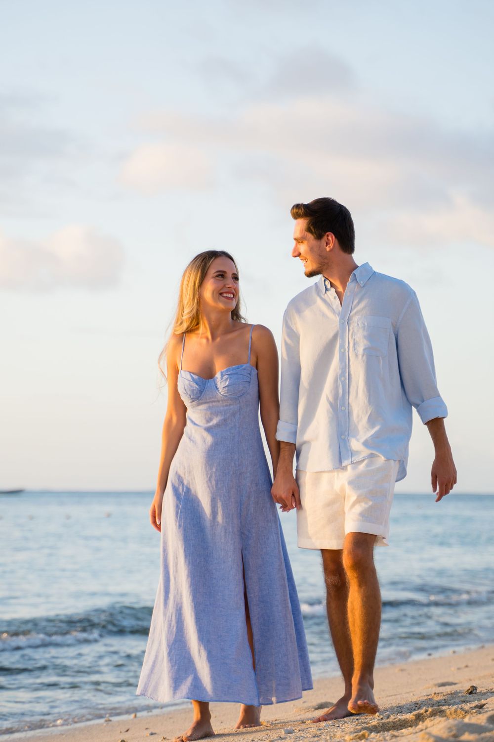 A couple in matching white and blue outfits walks along the shoreline.