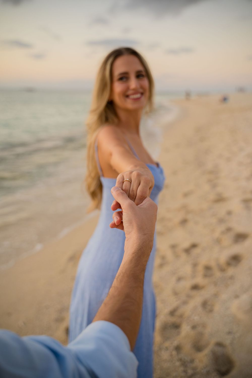 Woman in blue dress leading someone by the hand along a scenic beach at sunset.