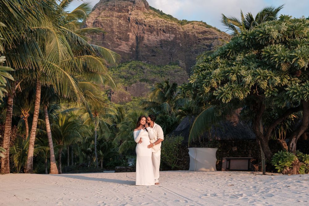 A couple poses on white sand beach with mountain and palm trees.