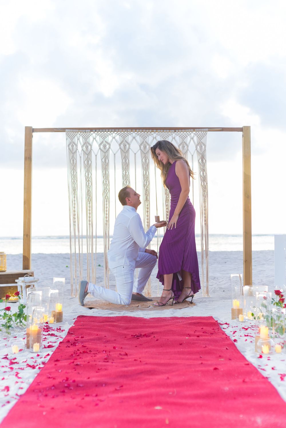 Romantic beach proposal setup with macrame arch, red carpet, and candles at sunset.