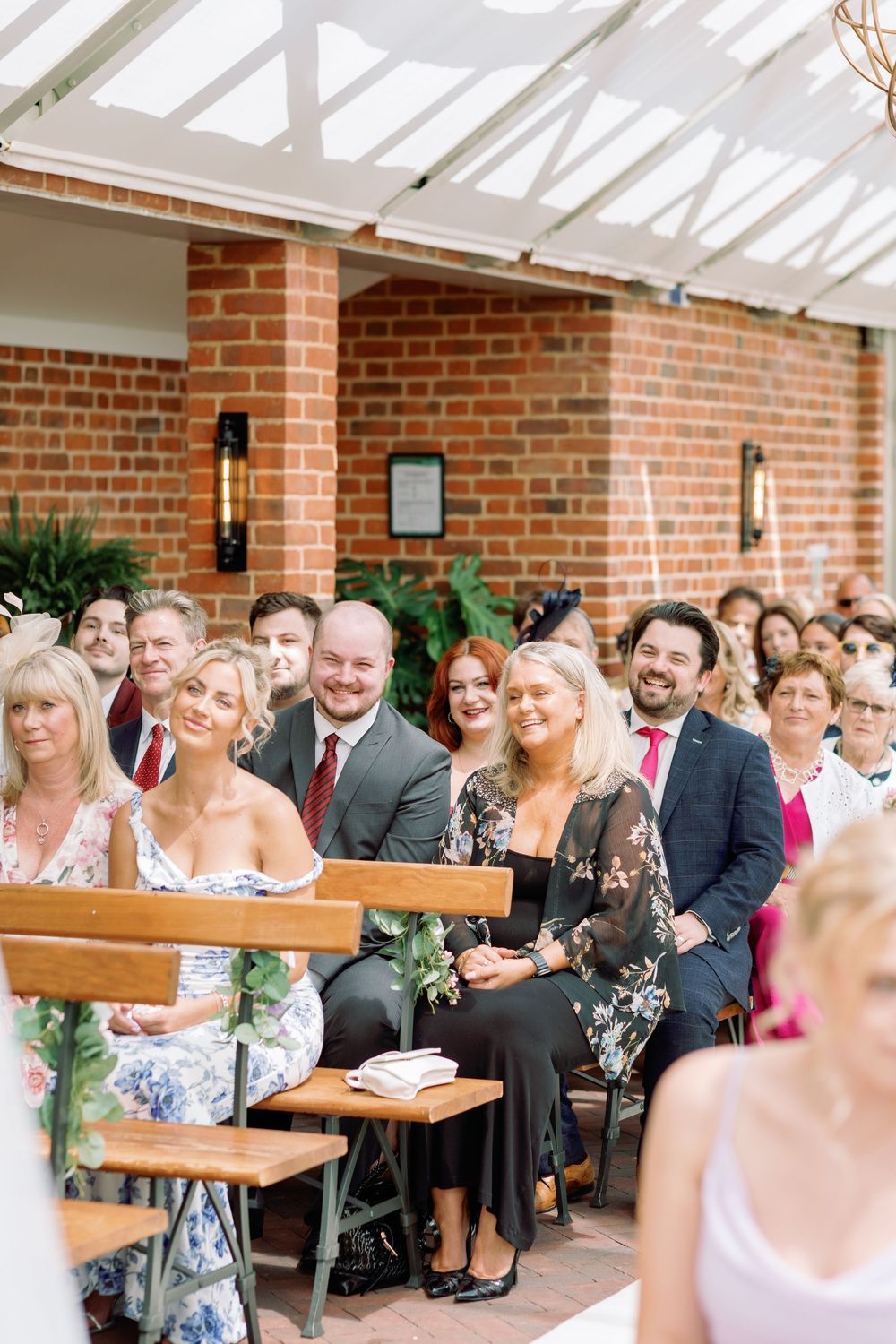 Wedding guests seated in rows on wooden benches at an outdoor ceremony under a covered brick patio.