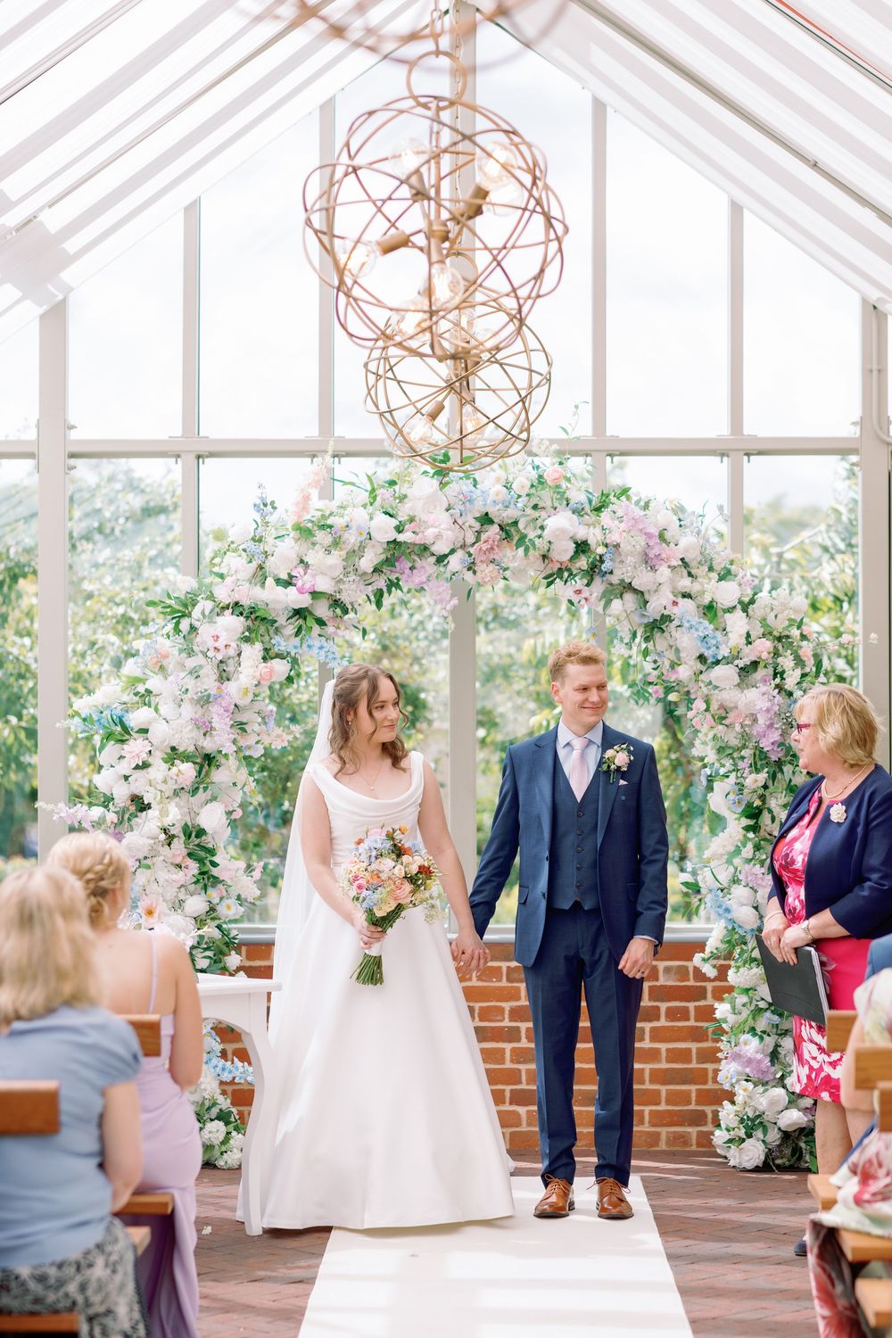Wedding ceremony in a conservatory decorated with elegant floral arrangements and copper chandeliers.