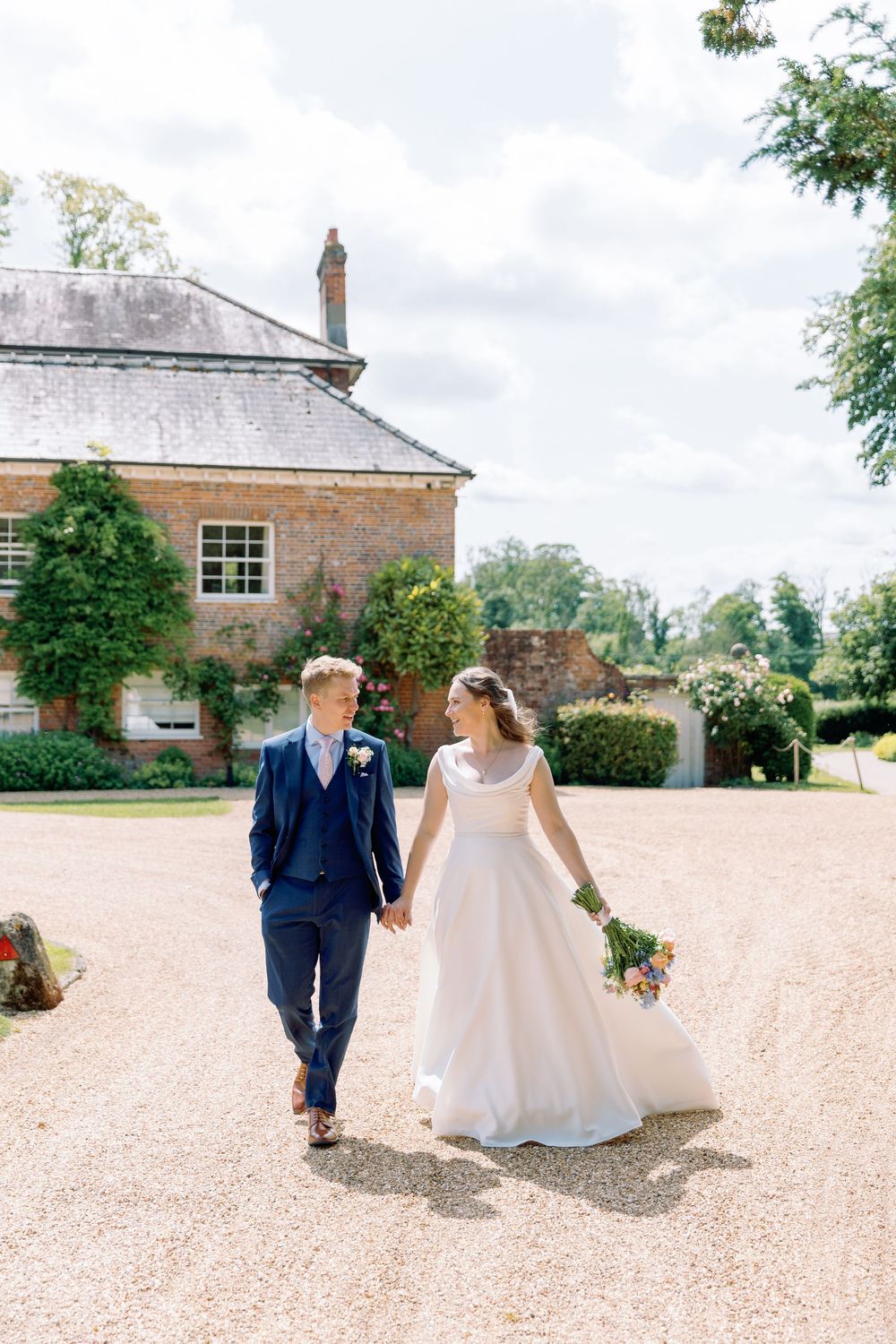 A couple walks hand in hand on a gravel path in front of a historic brick mansion on their wedding day.