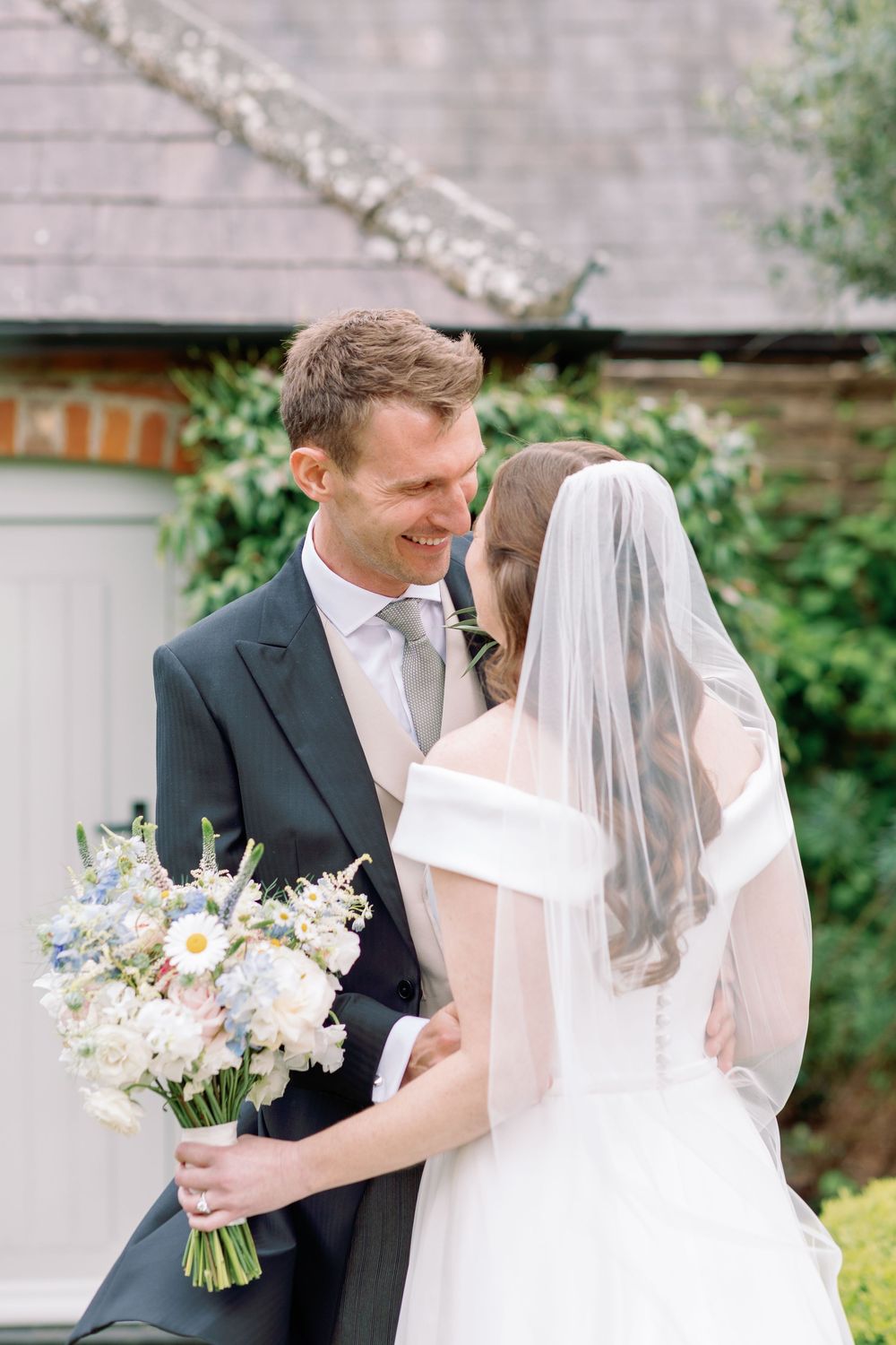 A tender wedding moment captured outdoors with a beautiful bouquet of wildflowers and daisies against a rustic backdrop.