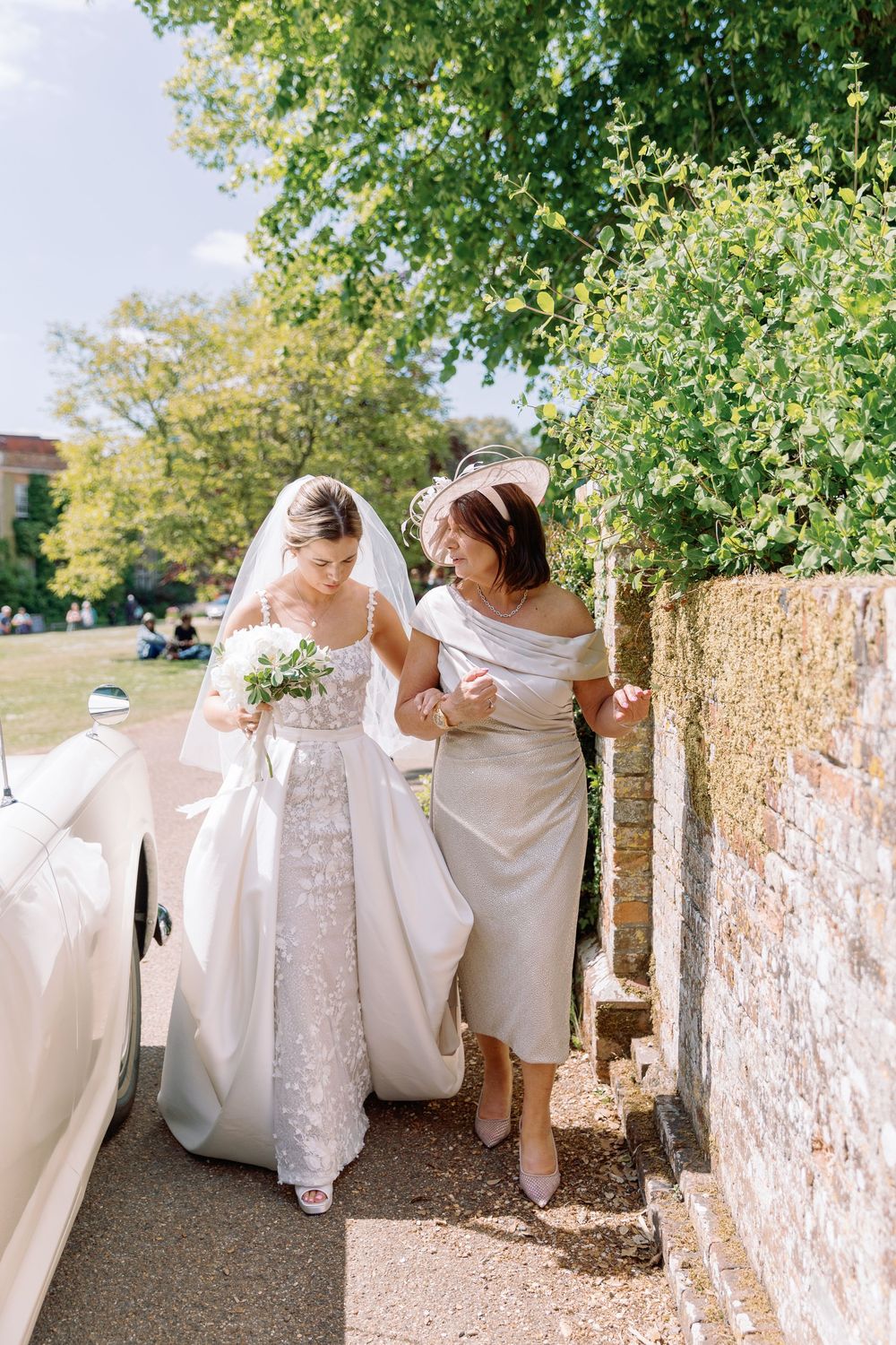 A wedding party in white dresses walks together along a stone wall path under green trees on a sunny day.