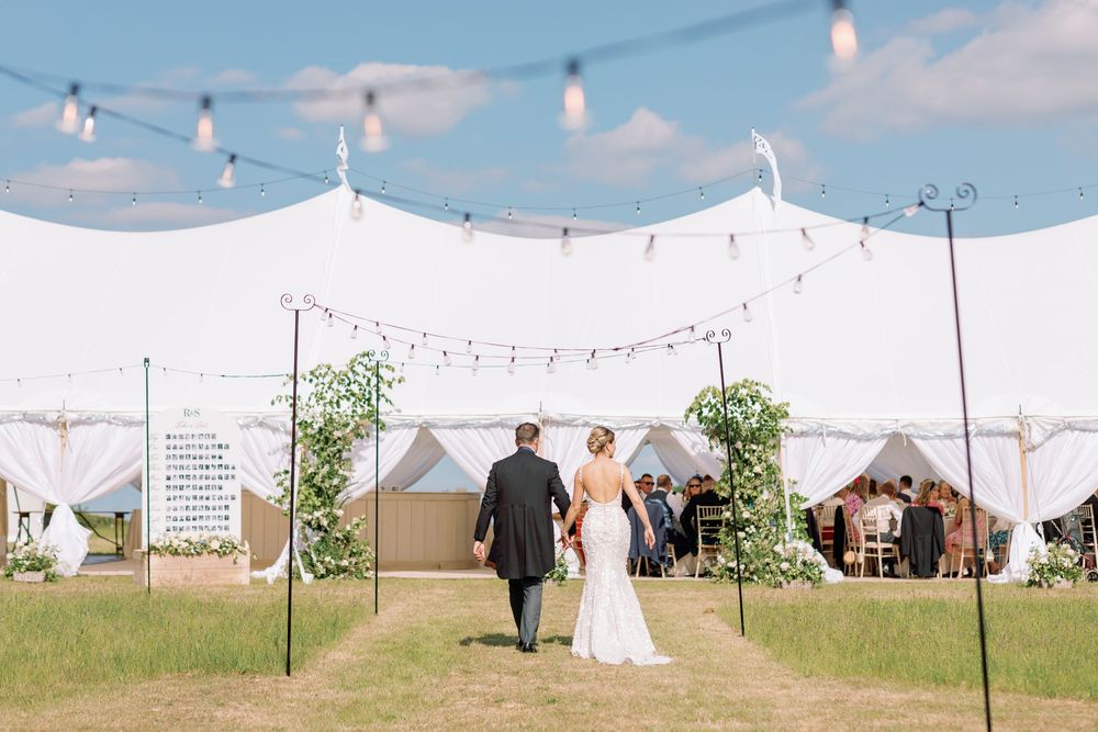 String lights hang over an elegant outdoor wedding reception setup with white tents and rolling hills in the background.