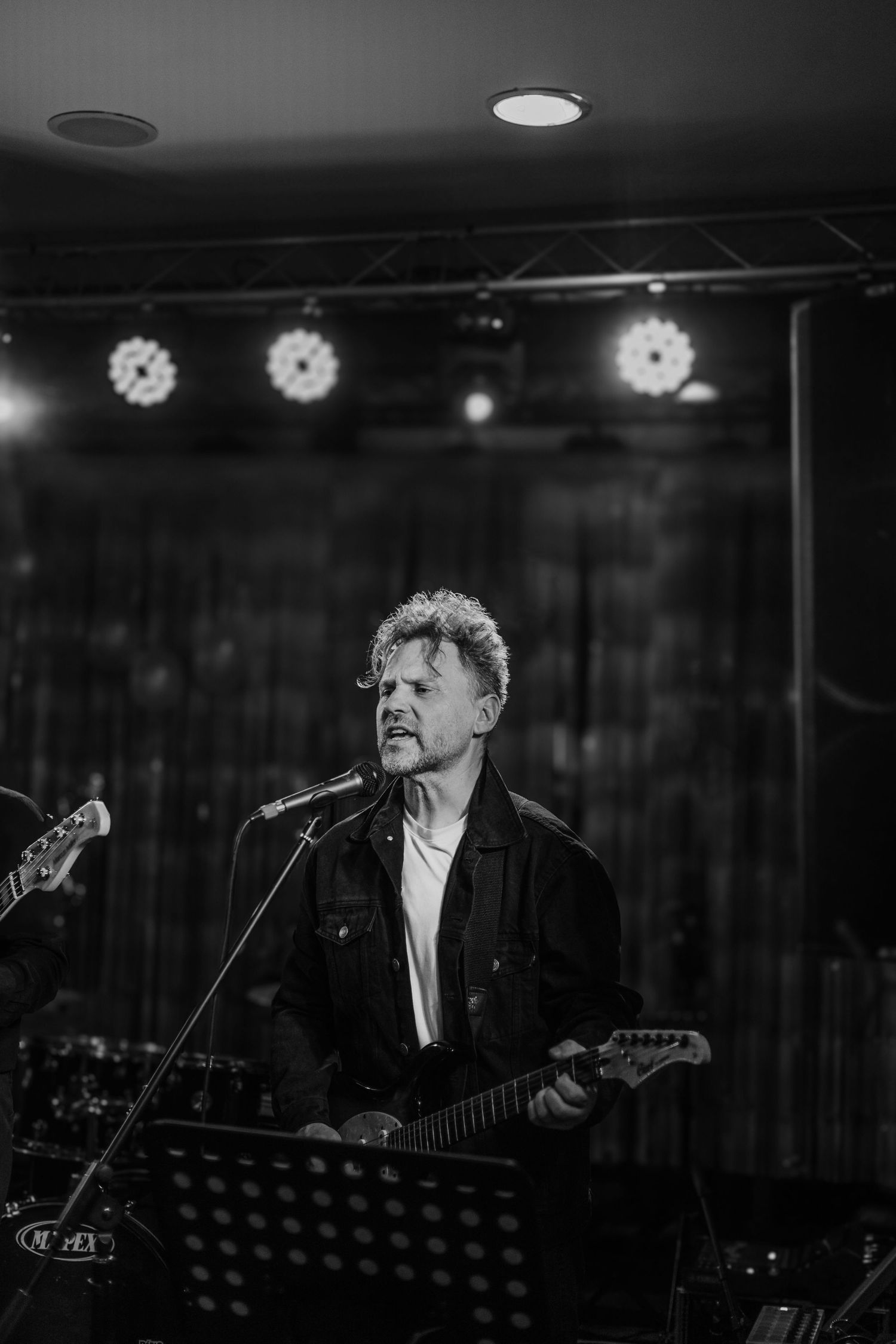 Detailný záber hudobníka s gitarou na pódiu, umelecká koncertná fotografia v čiernobielych tónoch.