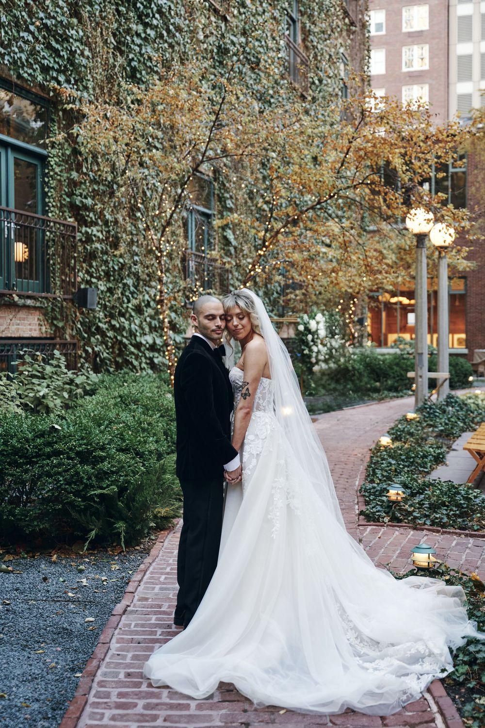 A romantic wedding couple poses on a brick pathway surrounded by ivy-covered walls and autumn foliage.